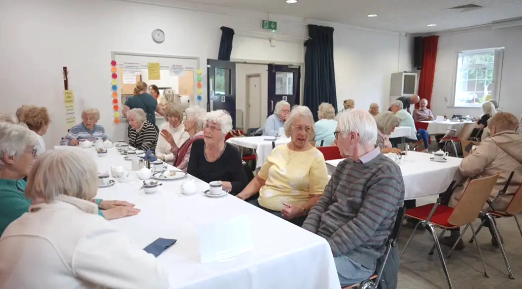 A lively social gathering in the Small Room, with community members seated at round tables enjoying tea and conversation.